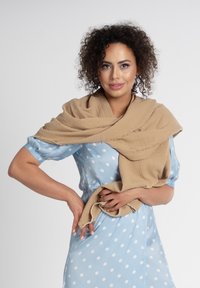 Beżowy, pleciony szal z teksturowanej tkaniny opadający na ramiona, trzymany w jednej ręce. Niebieska sukienka w kropki z krótkimi, bufiastymi rękawami.