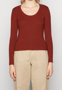 Rippenshirt mit langen Ärmeln in rostfarbener Farbe und Rundhalsausschnitt, kombiniert mit hellbeigen Hosen, was ein lässiges Outfit ergibt.