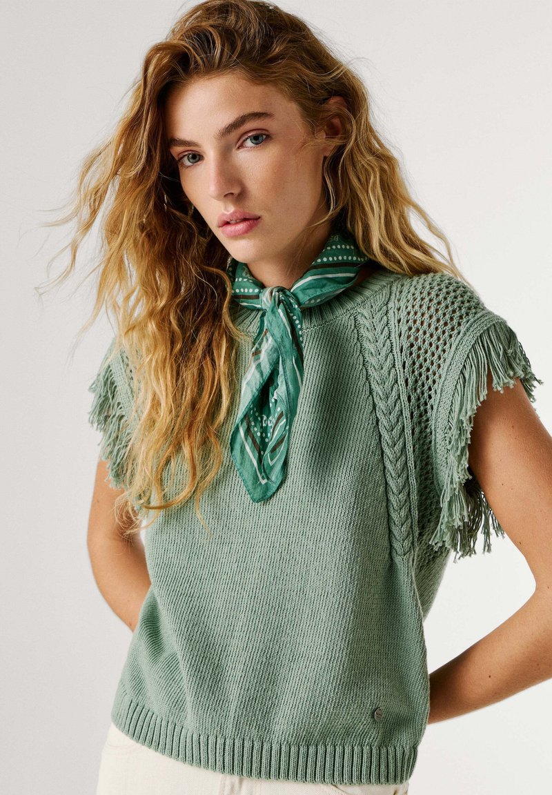 Junge Frau mit gewelltem blondem Haar, die ein salbeigrünes, gestricktes, ärmelloses Top mit Fransen und ein grünes, gemustertes Halstuch trägt.