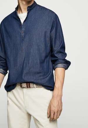 Homme portant une chemise en jean bleu foncé à manches longues retroussées sur un maillot blanc, un pantalon beige et une ceinture tressée.