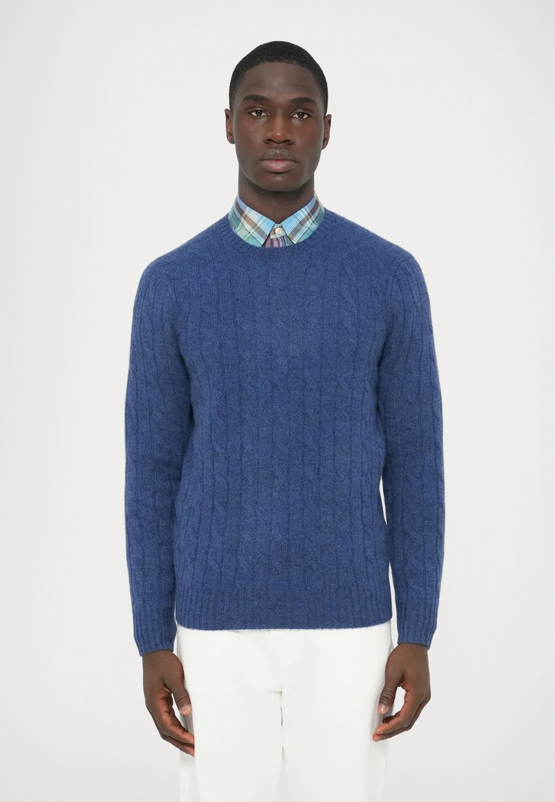 Kék, kötött pulóver puha anyagból, kerek nyakkivágással és hosszú ujjakkal, alatta kockás inggel párosítva.