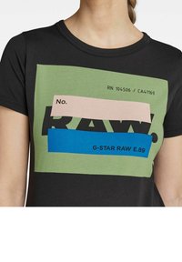 Kvinna som bär en svart t-shirt med gröna, rosa och blå grafiska block och texten "No. RAW" och "G-STAR RAW E.89" på framsidan.