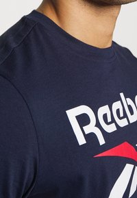 Marinblå bomulls-t-shirt med en vit Reebok-logotyp med röda och vita detaljer. Korta ärmar och rund halsringning, slät textur.