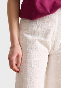 White embroidered pants featuring a square pattern with floral accents. The fabric has a textured finish. A hand with a gold bracelet is visible.