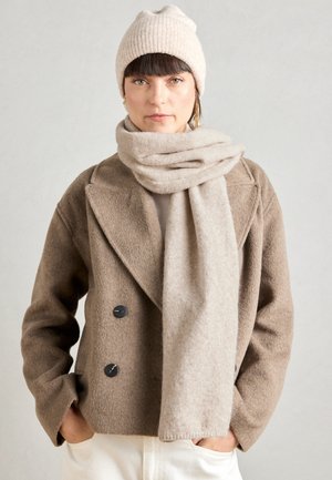 Beiger Wollmantel mit großen Revers und doppelten Knöpfen, kombiniert mit einem passenden beige Schal. Das Modell trägt eine gerippte beige Strickmütze.