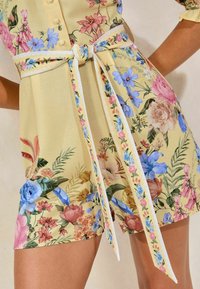 Floral-patterned dress in yellow fabric featuring pink, blue, and green flowers. Includes a white belt and ribbon accents for detailing.