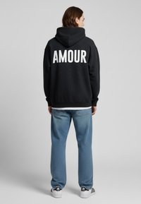 Felpa nera con "AMOUR" in grandi lettere bianche sulla schiena, abbinata a jeans azzurri chiari e sneakers in bianco e nero.