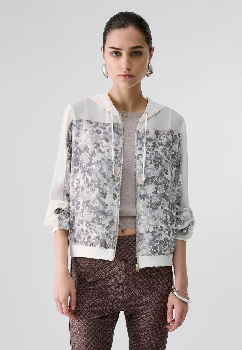 Jeune femme aux cheveux plaqués en arrière porte un sweat à capuche zippé blanc et gris à motif floral, un haut beige, un pantalon marron texturé et des boucles d'oreilles créoles argentées.