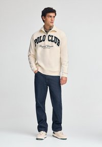 Felpa leggera color crema con la scritta "POLO CLUB" in blu scuro, abbinata a jeans blu scuro e sneakers beige con accenti verdi.