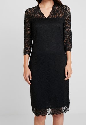 Vrouw draagt een zwarte knie-lange kanten jurk met driekwart doorzichtige mouwen en een gezoomde V-hals, staand tegen een effen achtergrond.