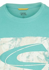 Lys turkis kortærmet t-shirt med rund hals, prydet med et tekstureret abstrakt hvidt print i midten og et gult mærke bagpå.