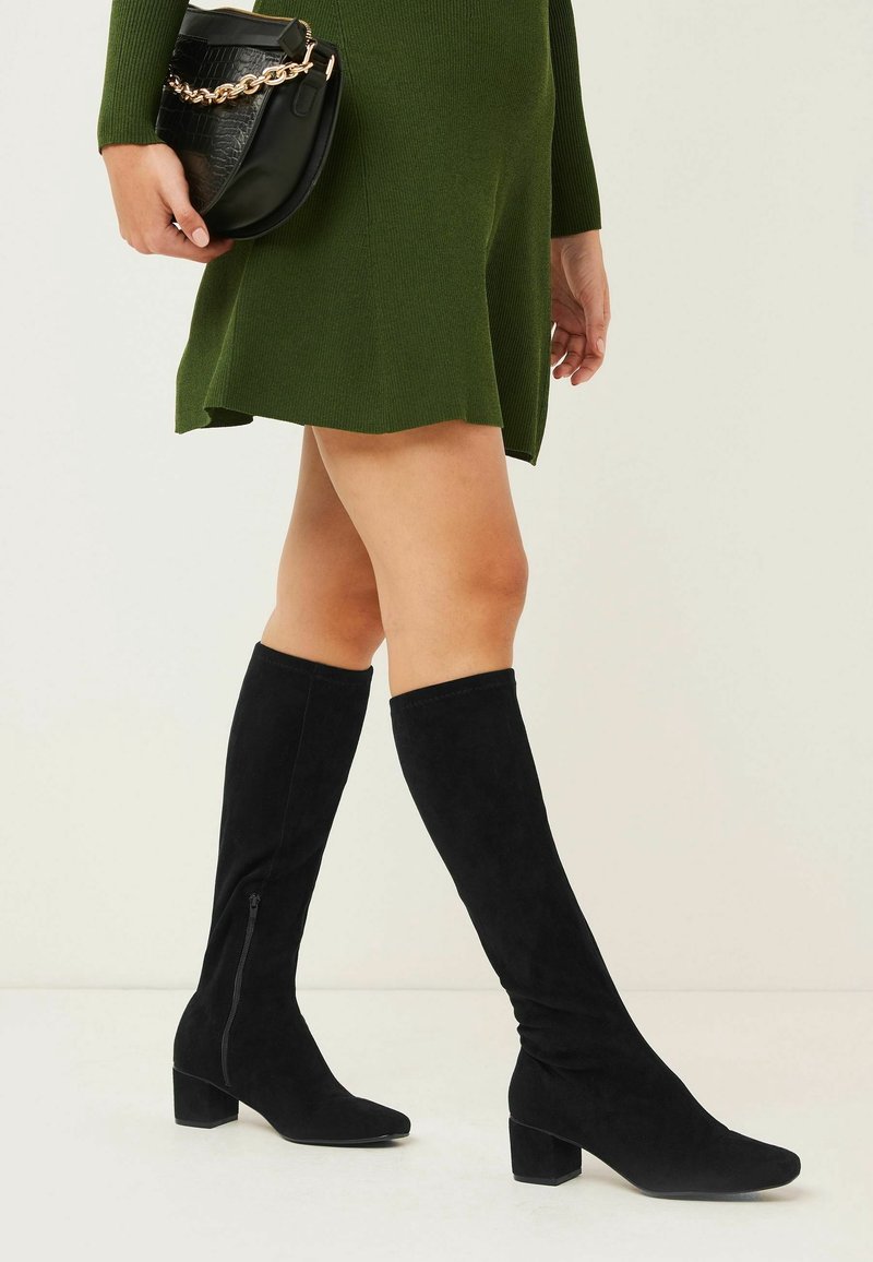 Bottes noires montantes en daim doux, avec un bout carré et un petit talon carré. Détail zippé sur le côté.