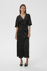 Melna satīna kleita ar V-veida izgriezumu un īs�ām piedurknēm. Ar grumbuļošanas detaļām viduklī un priekšējā šķēluma. Kombinēta ar melnām asām augstpapēžu kurpēm.