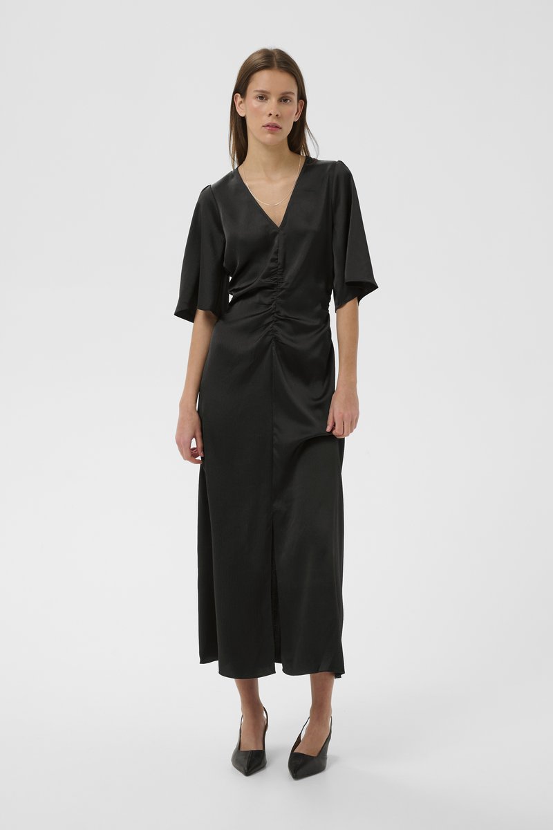 Melna satīna kleita ar V-veida izgriezumu un īsām piedurknēm. Ar grumbuļošanas detaļām viduklī un priekšējā šķēluma. Kombinēta ar melnām asām augstpapēžu kurpēm.