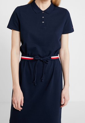 Mujer que lleva un vestido polo azul marino con mangas cortas y un cinturón de rayas rojo, blanco y azul atado a la cintura.