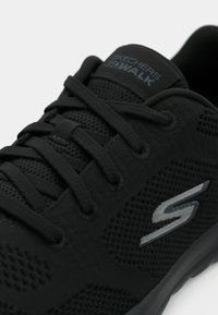 Gros plan sur une basket Skechers Go Walk noire avec tissu en mesh, lacets noirs et logo "S" argenté sur le côté.