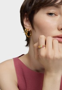 Złote kolczyki w kształcie kółek o gładkim wykończeniu, noszone z prążkowanym, różowym topem. Złoty pierścionek zdobią trzy kolorowe, przypominające kamienie akcenty.