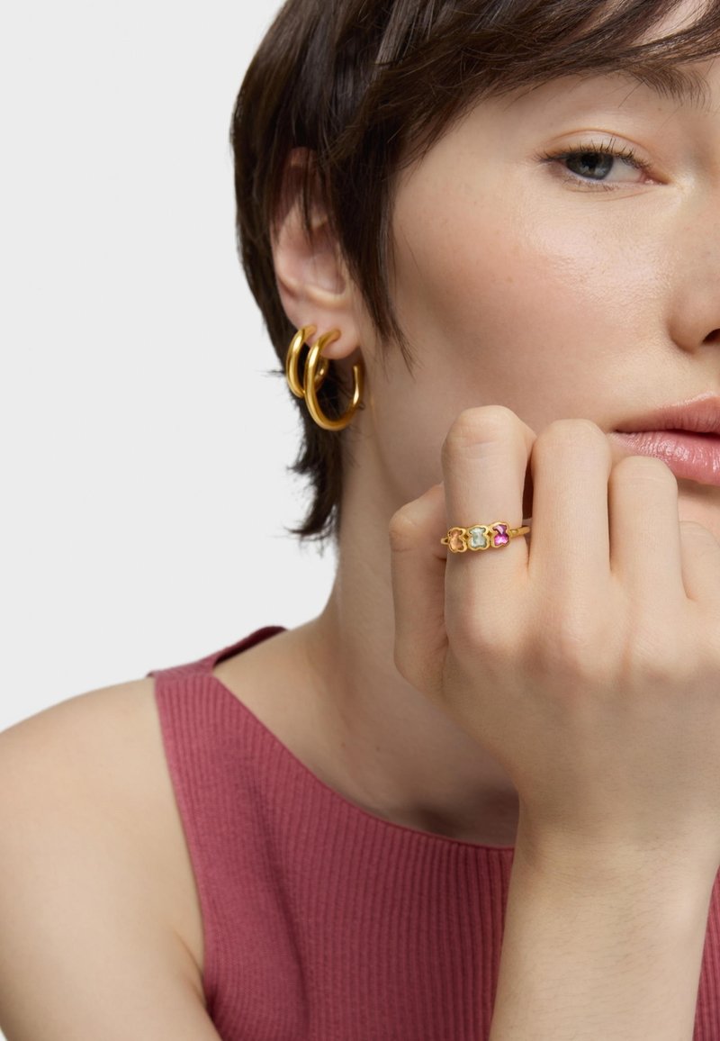 Złote kolczyki w kształcie kółek o gładkim wykończeniu, noszone z prążkowanym, różowym topem. Złoty pierścionek zdobią trzy kolorowe, przypominające kamienie akcenty.