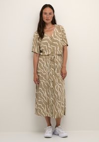 Beige, knielanges Kleid mit einem welligen weißen Muster, kurzen Ärmeln, Knopfleiste und verstellbarem Kordelzug in der Taille. Kombiniert mit weißen Sneakers.