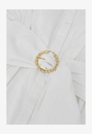 Valkoinen kangasvyö, joka on kiristetty kiertyneellä kultaisella pyöreällä soljella valkoisessa vaatetuksessa.