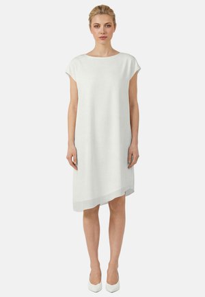 Kvinde står, iført en hvid knælængde kjole med korte ærmer, asymmetrisk kant og hvide spidse hæle.