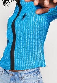 Blå randig zip-up topp med metallisk tråd, med en svart dragkedja och logodetalj, bärs tillsammans med ljus tvättade jeans.