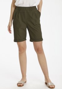 Pantaloni corti in verde oliva, sartoriali, con una cintura larga, pieghe e risvolti. Presentano due tasche laterali. Indossati con sandali a scivolo beige.