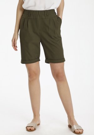 Pantaloni corti in verde oliva, sartoriali, con una cintura larga, pieghe e risvolti. Presentano due tasche laterali. Indossati con sandali a scivolo beige.