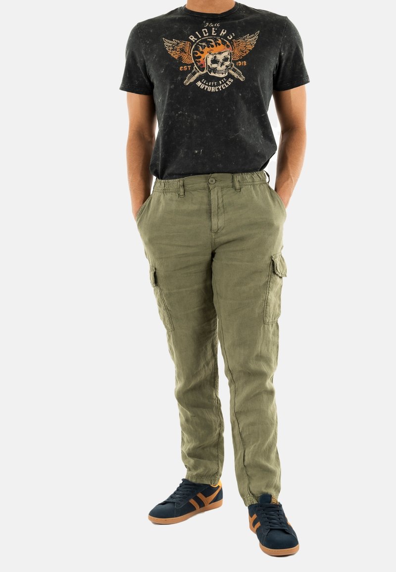 Pantaloni cargo verde oliva con tasche laterali, abbinati a una maglietta nera con grafica che presenta un design di teschio. Le scarpe sono blu navy con dettagli arancioni.