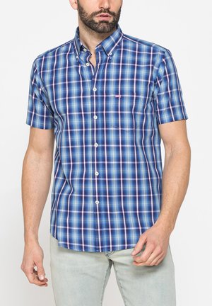 Uomo con barba che indossa una camicia a quadri blu e rosa a maniche corte e pantaloni grigio chiaro, in piedi su sfondo bianco.