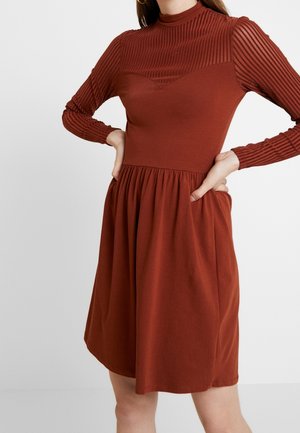 Femme portant une robe couleur rouille avec des manches longues côtelées et une jupe froncée, les mains posées sur les hanches sur un fond uni.
