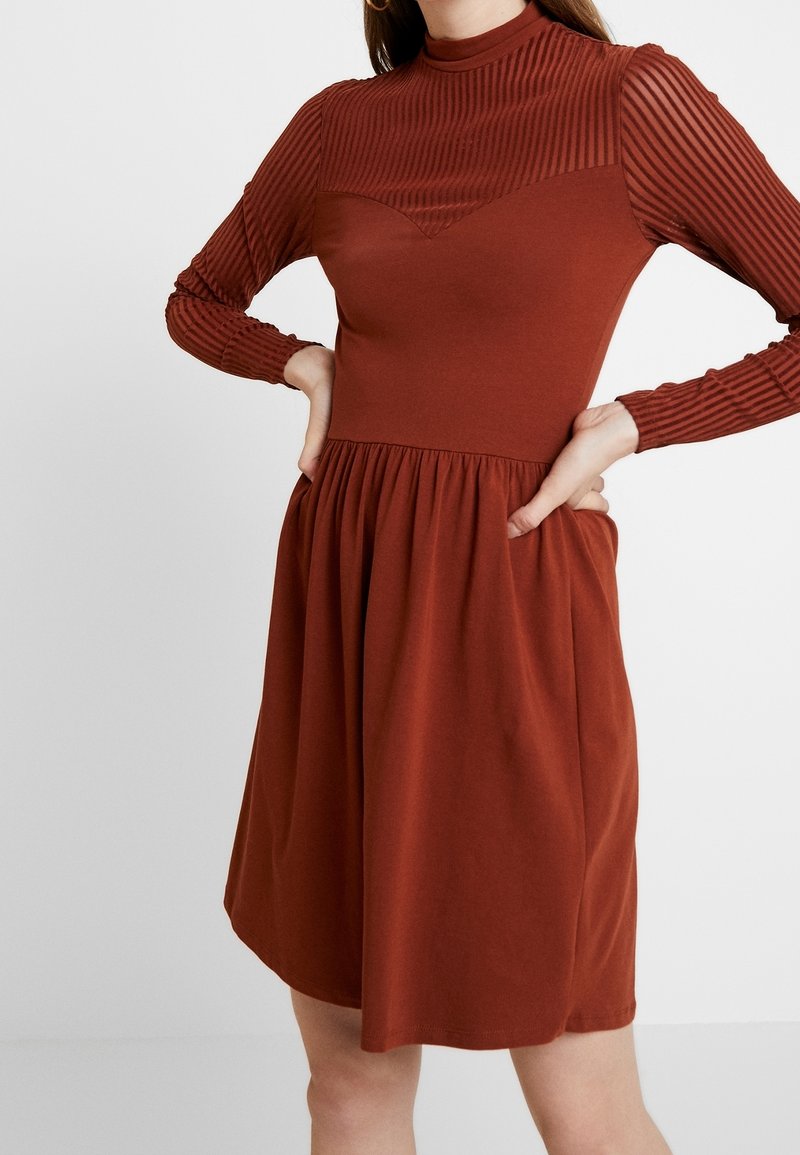 Femme portant une robe couleur rouille avec des manches longues côtelées et une jupe froncée, les mains posées sur les hanches sur un fond uni.