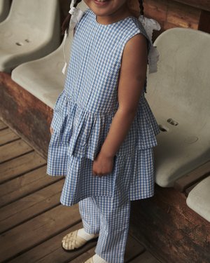 Ung pige med flettet hår iført en blå og hvid mønstret kjole og bukser, stående på trægulv ved siden af plastikstole.