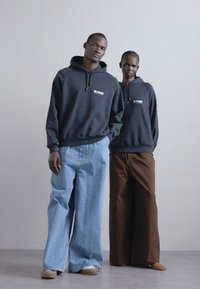 Deux modèles portant des sweats à capuche, l'un en gris foncé avec un logo et l'autre en marron, assortis à des jeans en denim à jambes larges. Les textures lisses dominent.