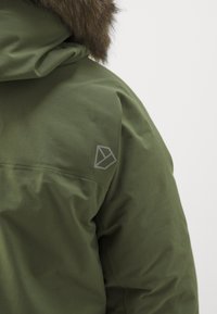 Parka verde oliva con cappuccio foderato in pelliccia. Presenta un logo geometrico sulla spalla e una finitura in tessuto liscia e testurizzata.