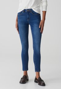 Jeans in denim blu scuro con una vestibilità slim, vita media, cuciture in contrasto e orlo alla caviglia, abbinati a mocassini scuri.