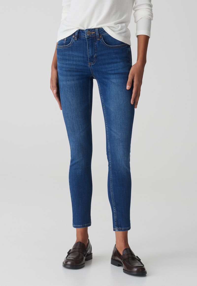 Jeans in denim blu scuro con una vestibilità slim, vita media, cuciture in contrasto e orlo alla caviglia, abbinati a mocassini scuri.