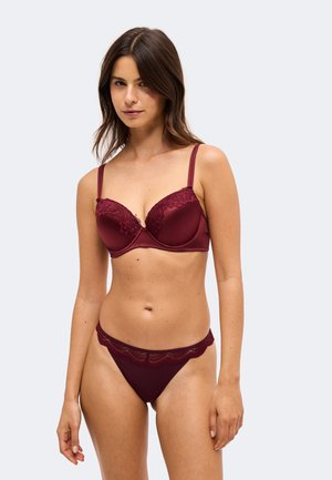 Femme aux cheveux bruns portant un soutien-gorge bordeaux orné de dentelle et une culotte assortie sur un fond clair uni.