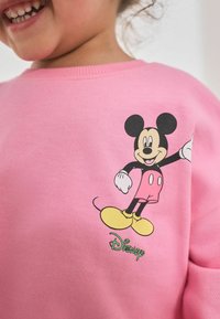 Pulover roz cu guler rotund și manșete cu nervuri, având un grafic cu Mickey Mouse negru și galben și textul "Disney" în verde.