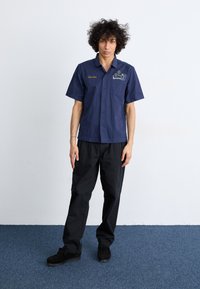 Marineblauw overhemd met korte mouwen met geborduurd bloemenpatroon en de naam "Jarvice," gecombineerd met zwarte wijde broek en zwarte schoenen.