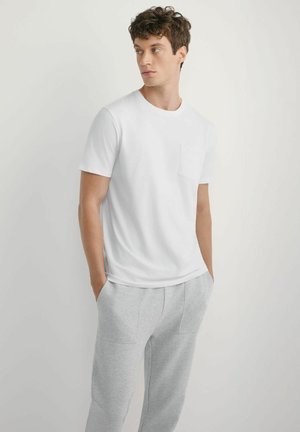 Młody mężczyzna z kręconymi włosami, ubrany w gładką białą krótką koszulkę i jasnoszare spodnie dresowe, stojący z rękami w kieszeniach.