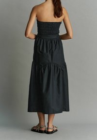 Musta olkaimeton maksi-mekko, jossa on smokattu yläosa ja kerroksittainen helma. Mekossa on pehmeä puuvillamateriaali ja röyhelöreuna, yhdistettynä tasapohjaisiin sandaaleihin.