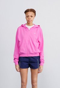 Felpa rosa con zip e cappuccio, realizzata in materiale morbido, con un piccolo logo bianco sul petto. Indossata su pantaloni corti scuri con dettagli laterali.