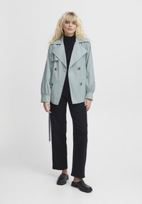 Hele sinine kahekordne trench coat suurte kraedega, kogutud varrukatega ja kahe ees oleva taskuga, koos mustade pükste ja kingadega.