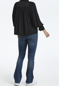 Svart, lätt blus med lång ärm och rynkad detalj i ryggen, matchad med blå flare jeans och vita sneakers.