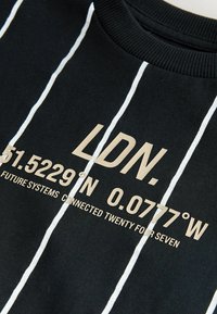 Felpa nera con strisce verticali bianche. Il testo recita "LDN. 51.5229°N 0.0777°W FUTURE SYSTEMS CONNECTED TWENTY FOUR SEVEN" in oro.