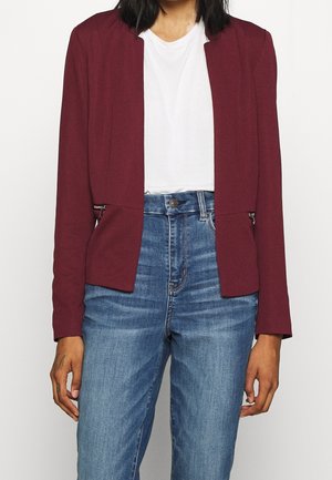 Henkilö, joka on pukeutunut valkoiseen t-paitaan, viininpunaisiin blazer-housuihin, joissa on vetoketjulliset taskut, ja korkeavyötäröisiin sinisiin farkkuihin vaaleaa taustaa vasten.