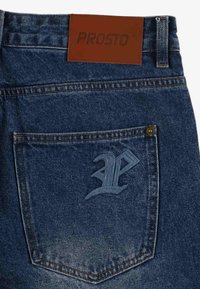 Jeans dżinsowe z brązową skórzaną naszywką w pasie oznaczoną "PROSTO". Posiada tylną kieszeń z wytłoczonym logo "P". Ciemnoniebieski kolor.