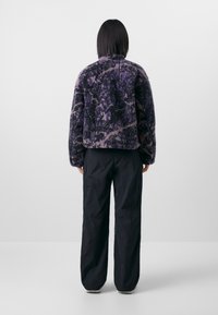 Giacca in pelliccia sintetica con motivo astratto viola e nero, caratterizzata da un colletto alto e una vestibilità comoda. Abbinata a pantaloni neri a vestibilità ampia.