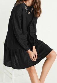 Mujer sentada en un taburete blanco, vestida con un vestido negro de manga larga con dobladillo fruncido y un gran collar de eslabones ovalados de plata.